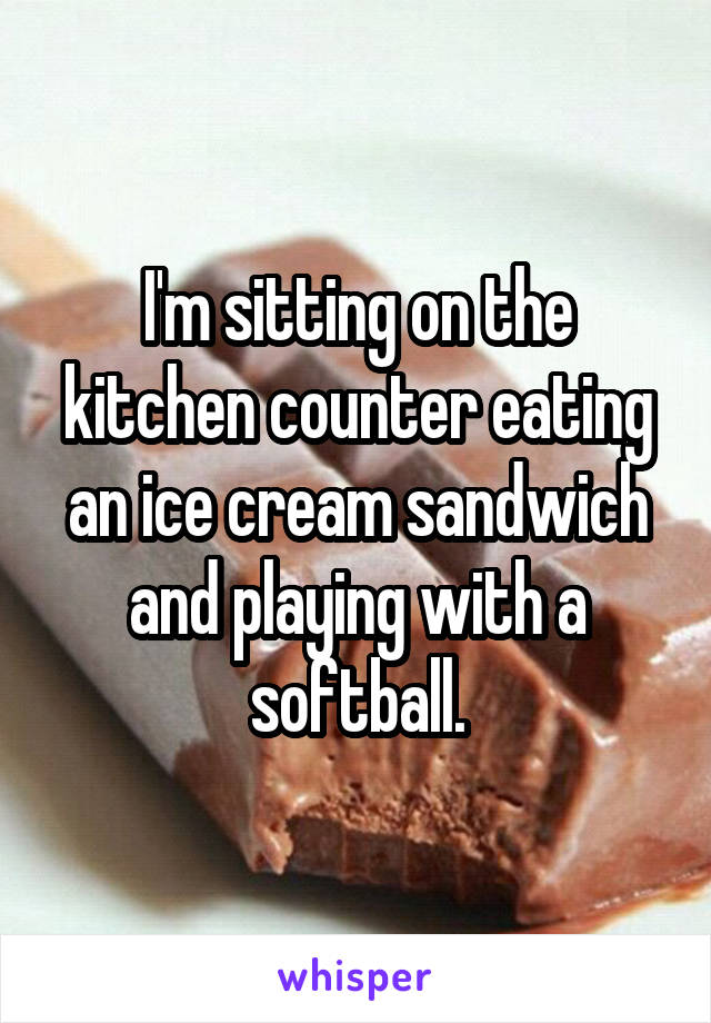 I'm sitting on the kitchen counter eating an ice cream sandwich and playing with a softball.